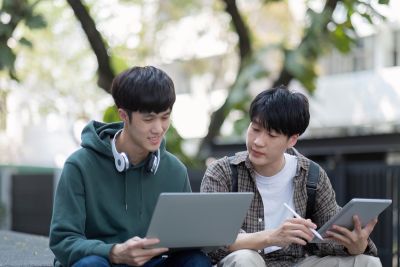 Two high school students are sitting on some outdoor steps in a cityscape looking at the same laptop
