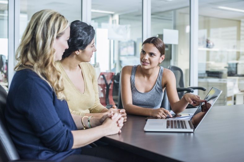 Image of an Aboriginal woman talking to female colleagues in modern office and pointing to laptop computer.