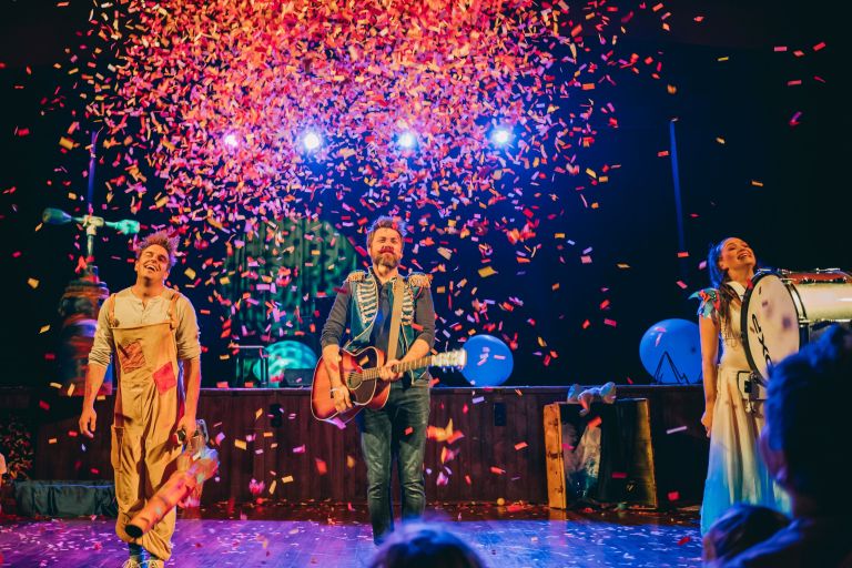 Three people on a stage holding instruments with confetti falling down on them