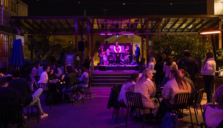 Groups of people sitting around tables at an outdoor venue at night, watching a band perform.