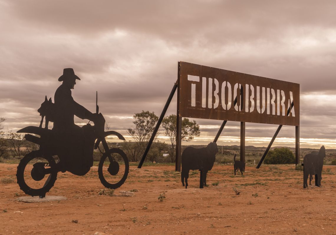 Tibooburra road sign with sculptures of a person riding a motorcycle, sheep and a dog.