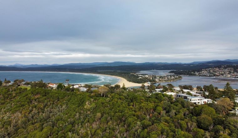 Looking down from forested hill over an ocean inlet with lake surounded by houses and hills in the background.