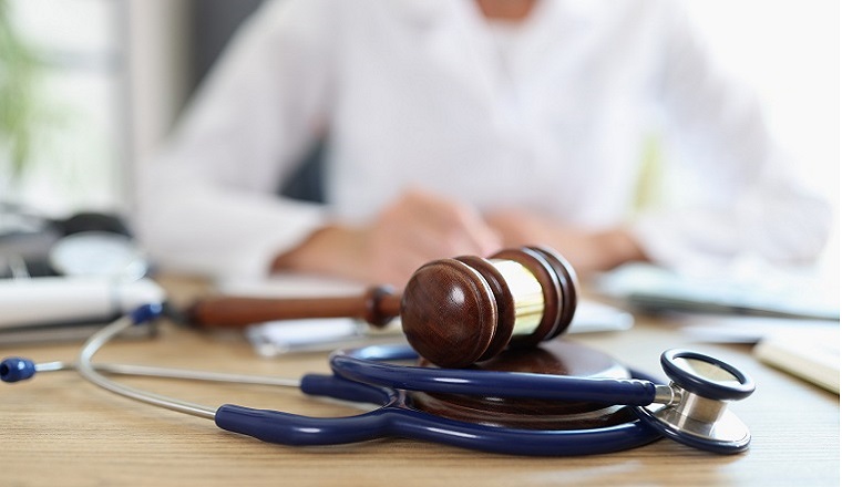 A wooden gavel and a navy stethoscope rest on a wooden desk. In the background, a person in a white lab coat is seated, appearing engaged in work.