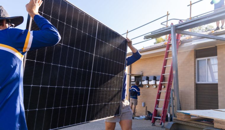 Two people carrying a solar panel in front of a house under construction.