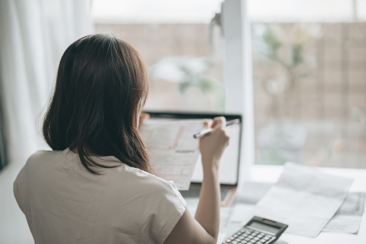 Woman with back turned holding pen and form in front of window, with calculator and laptop visible on desk in front of her.