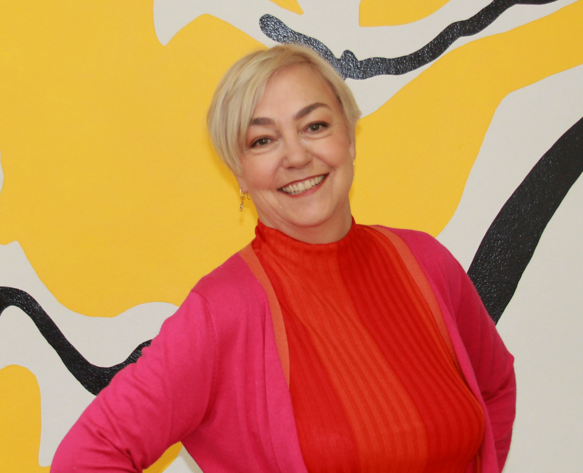 A woman is standing in front of a yellow artwork. She is smiling. She is wearing a pink and orange outfit.