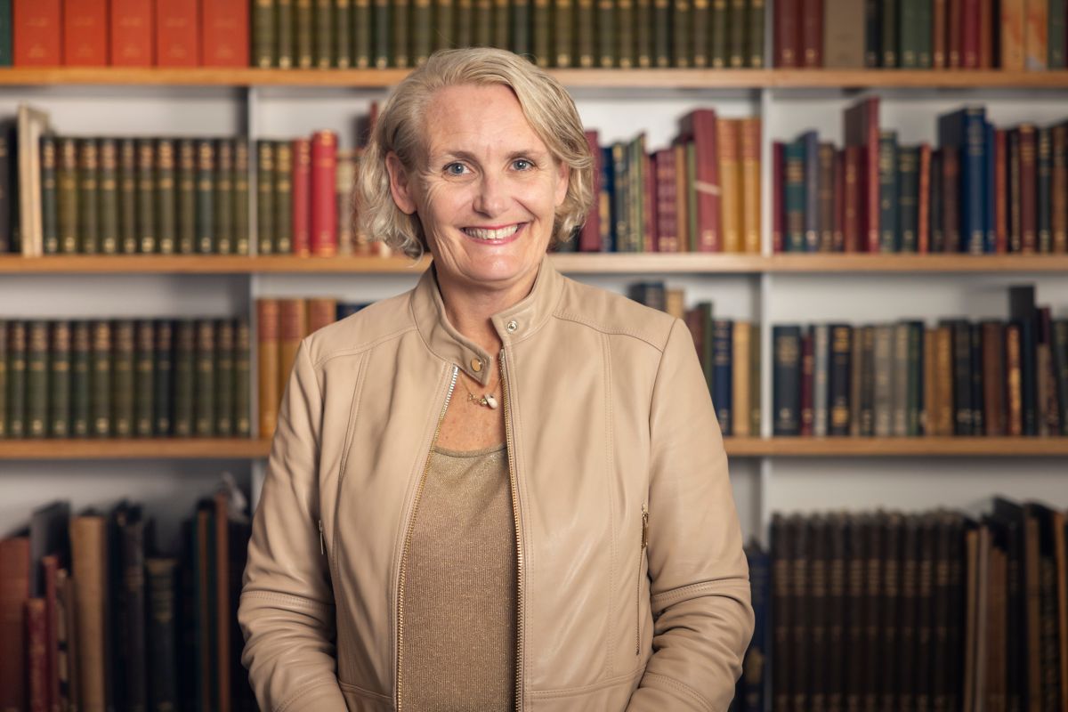 A woman is standing in front of a bookshelf. She is wearing a tan blouse and jacket. She is smiling. 