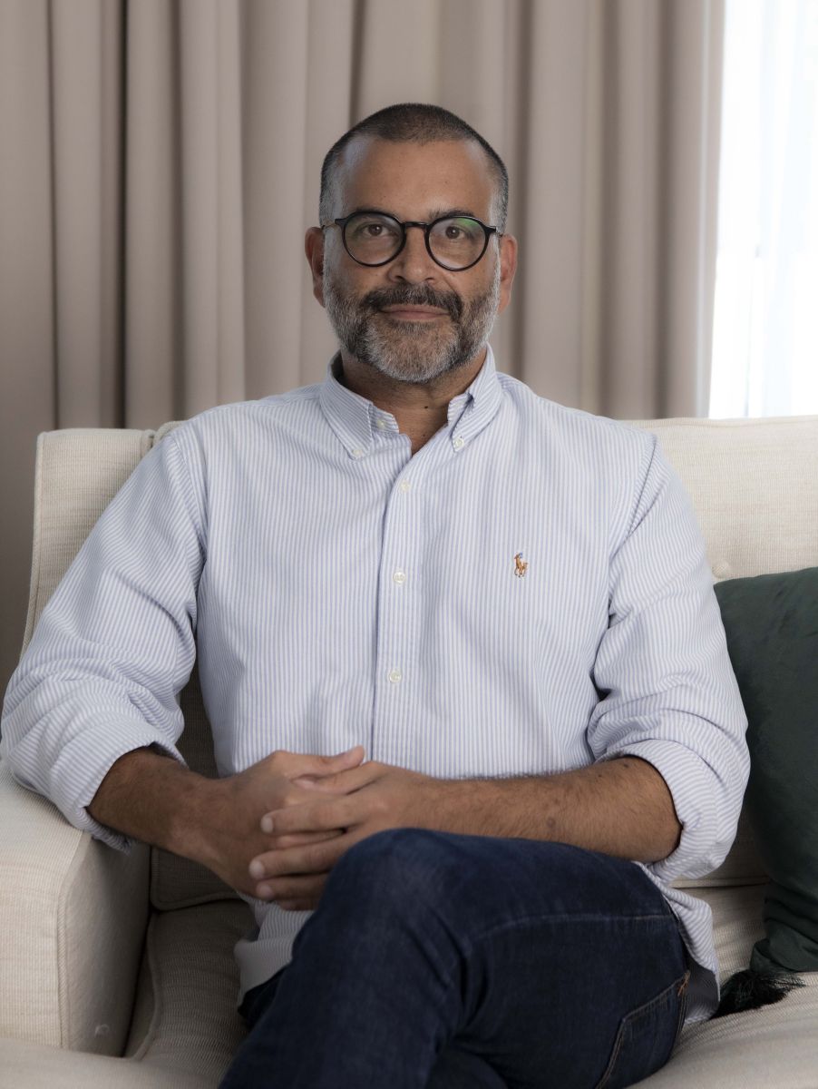 A man is sitting on a couch and looking at the camera. He is wearing glasses.