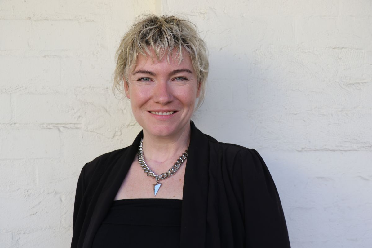 A woman is staring at the camera and smiling. She is standing in front of a white brick wall. She is wearing a black outfit. She is also wearing a necklace.