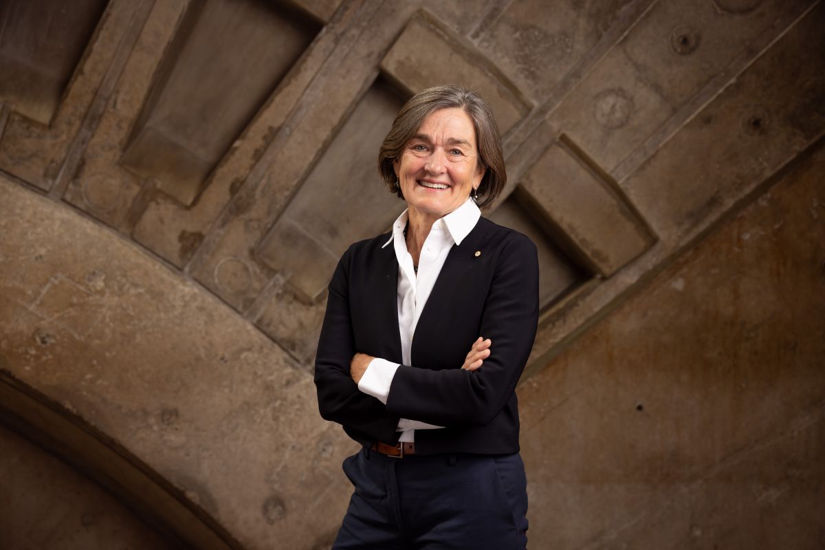 A woman is standing and smiling at the camera. She has her arms crossed. She is wearing navy blue pants, a white blouse and black tailored jacket.