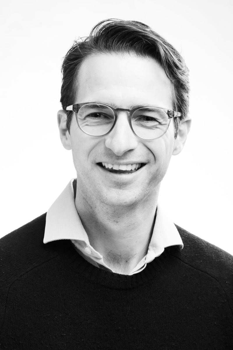 A man is looking at the camera and smiling. He is wearing a shirt and jumper. He is also wearing glasses.