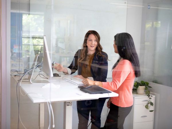 Two women stand at a computer facing each other as one woman points at the computer screen