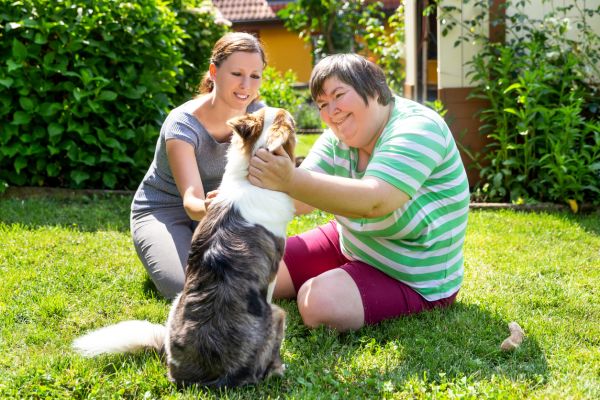 Woman with intellectual disability sitting on grass with carer patting dog