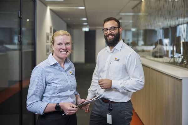 Woman with blonde hair wearing a blue checkered shirt and holding some paper, standing next to a man with brown hair, a brown beard and white shirt. Both are standing up while smiling at the camera.