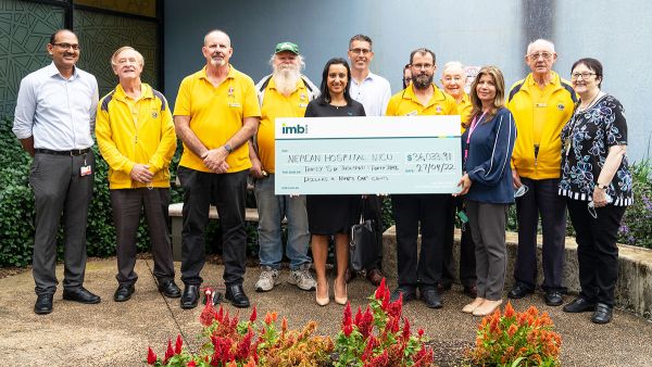 A group of adults hold up a giant cheque donation while standing outside a health facility