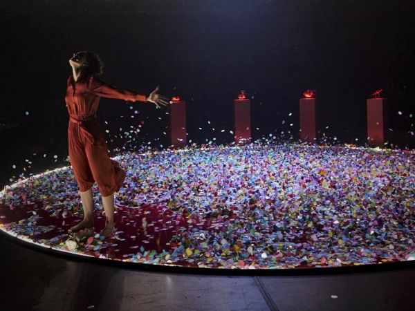 Woman standing arms outstretched against breeze on stage, with coloured paper flying behind