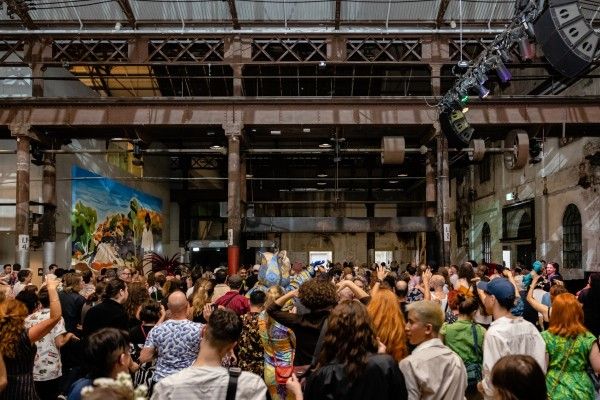 A crowd looking towards a stage with industrial lighting hanging over them. 