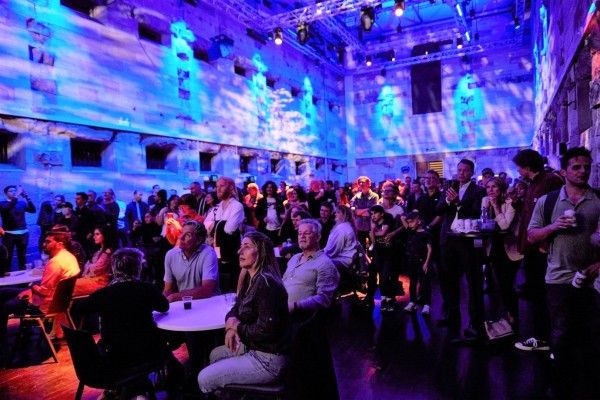 A large crowd sitting inside a building and high tables surrounded by blue lighting 