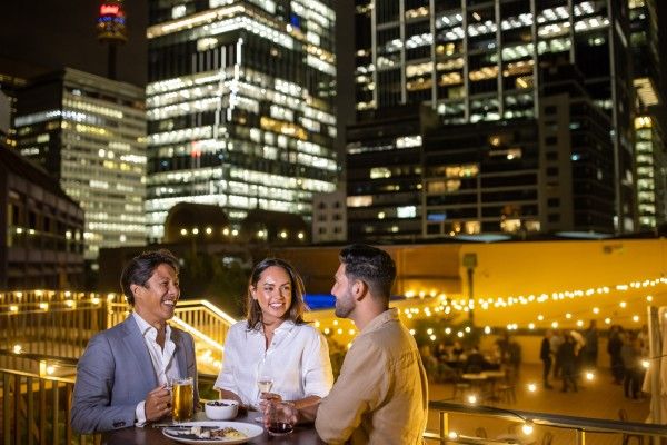 3 people standing in front of brightly lit buildings at nighttime, with a table of food and drinks