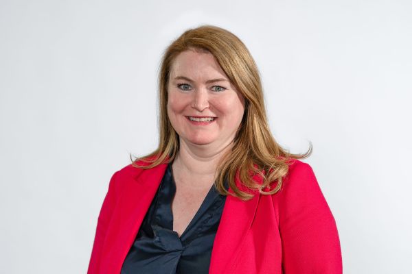 A person with  red shoulder length hair smiles at the camera and wears a red suit jacket and black blouse