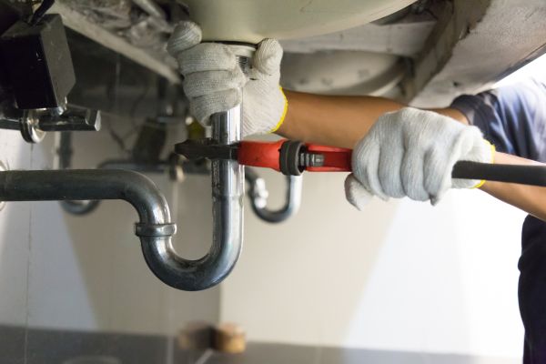 Hands holding wrench attached to plumbing pipes under sink.