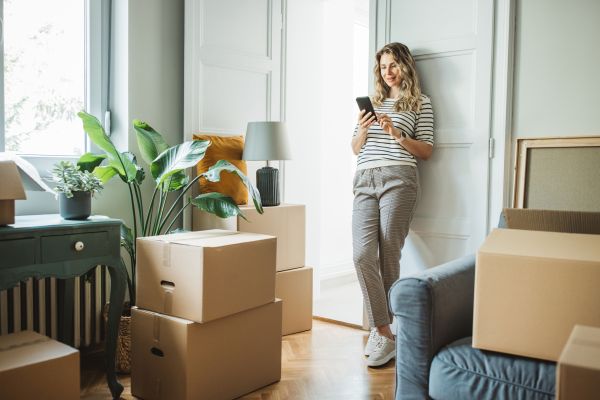 Woman leaning on wall looking at mobile phone, with cardboard boxes, furniture and pot plant.