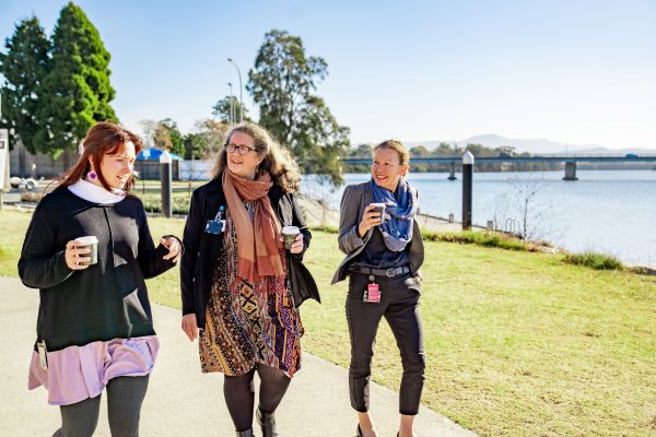 Three women talking and walking outdoors, each of them are holding a takeaway coffee.   