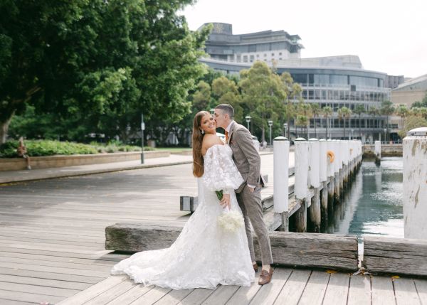a bride and groom post for the camera. They are stand on a bayside wharf, behind them you can see the blue water and the green of the trees,