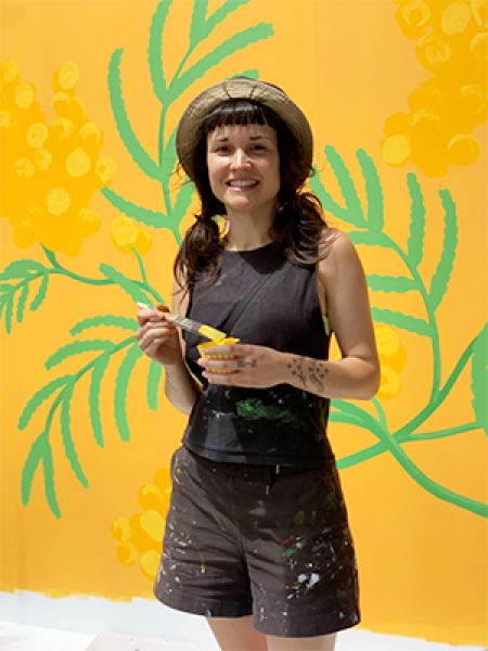Pilar Basa holds a paintbrush in front of her vibrant mural of local Parramatta wattle.