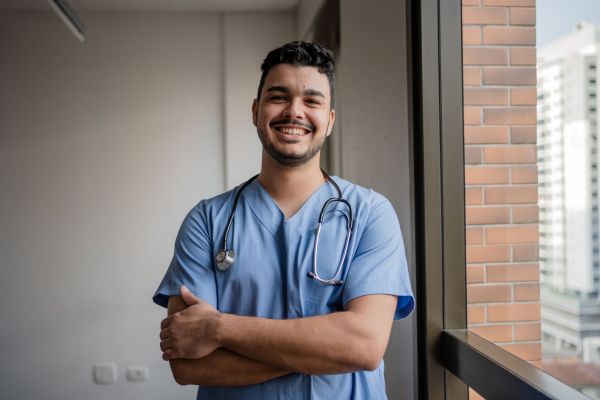 A male nurse stands in his office, smiling with arms folded.