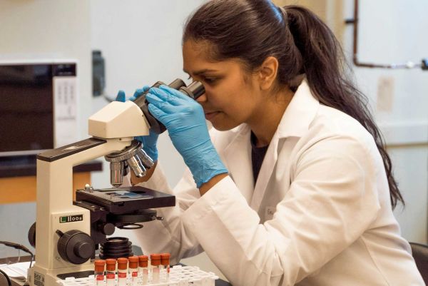 A woman in a white lab coat, looking through a microscope