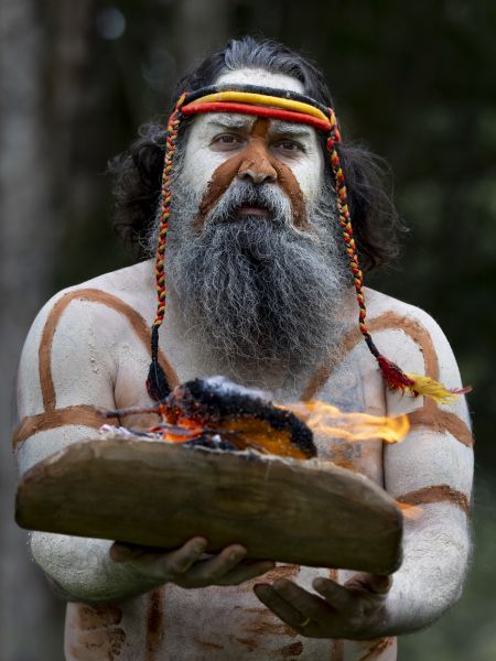 An aboriginal man in traditional paint holding up a burning pile of leaves and wood for a smoke ceremony. 