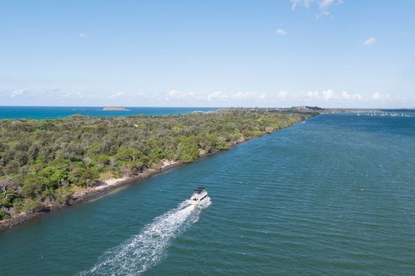 A white boat with a white wake behind it jets across deep blue water. To its left is a narrow peninsula covered in trees with water on the other side