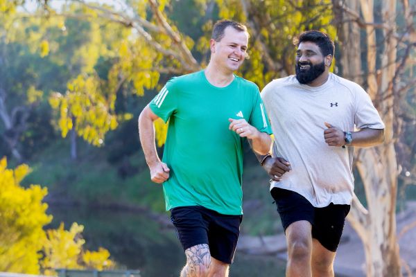 Two people in running gear - one in a bright green shirt and the other in white - jog along a path with trees in the background