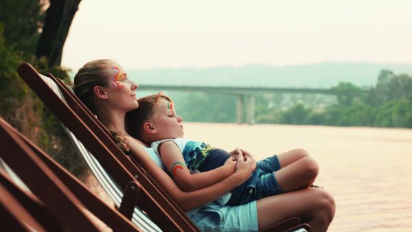 A woman and a child relaxing on deck chairs by a river, gazing peacefully at the water, with a distant bridge and soft, hazy landscape in the background.