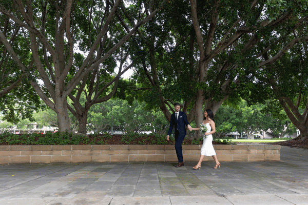 A bride and groom hold hands as they walk in front of a row of trees outside the Pyrmont Wedding Registry.