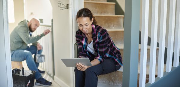 A woman sits on unfinished stairs looking at a tablet while a man paints the house in the background.