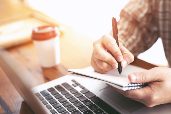 Person writing on notepad next to a laptop