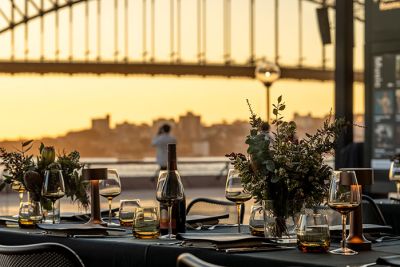 A dining table styled for an event with wine glasses and native flowers, with the Sydney Harbour Bridge in the background.