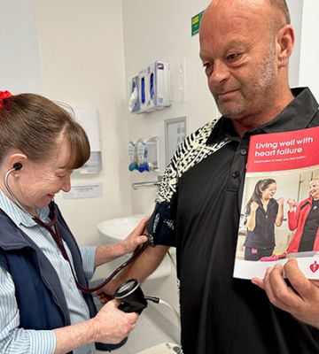 Patient holding an information brochure about heart failure, while having his blood pressure reading taken by a smiling nurse.