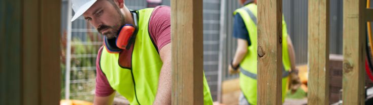 Male bricklayer cementing bricks. Female engineer is working in background. They are wearing reflective clothing at a building site.