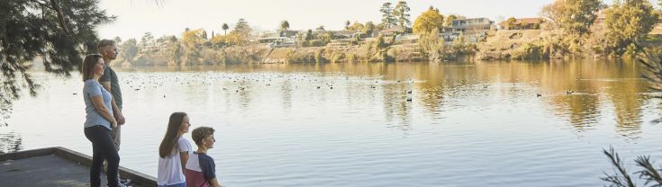 Two adults stand on a pier overlooking the River on a sunny day in Penrith. Their children are sitting on their pier with their feet dangling over the edge. There are birds on the river.