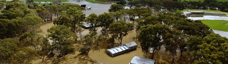 Aerial view of a flooded farm showing a home, sheds and trees sitting in brown water, with green hills in the background