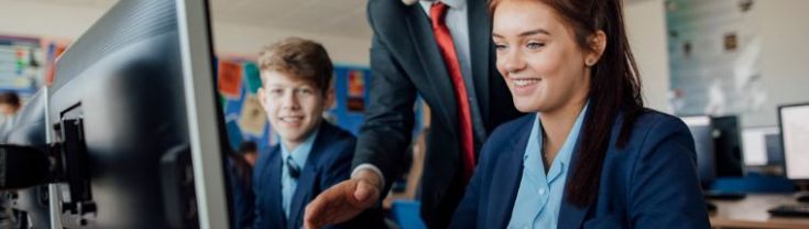 Two pupils, a male and a female, are sitting behind computers receiving help from a male teacher during a lesson.