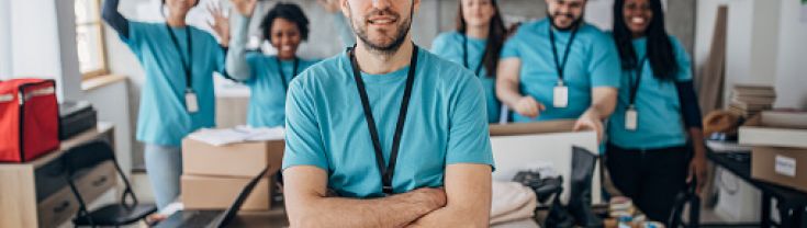 Man in volunteer tshirt stands with folded arms in front of a team of volunteers packing boxes in background