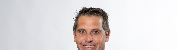 A person with brown hair wears a blue checked suit with a blue striped business shirt and dark blue tie. He smiles at the camera