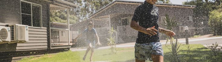 Children run through a water sprinkler outdoors on a lawn, next to a house.