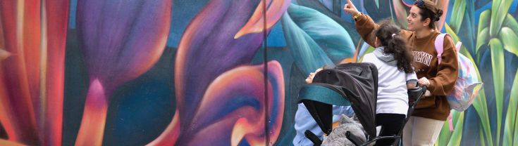 A woman pushes a pram with a child inside, while looking at a painting on an outdoor wall in Liverpool.