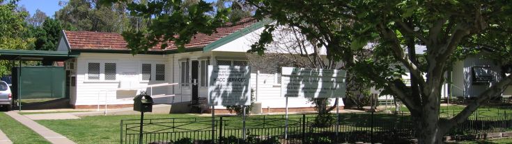 Main entrance to the Moulamein Community Health Centre. The centre's small driveway sits to the left side of the image. A large tree is sited on the verge outside the centre's fenced area. Two white signboards sit on the main lawn of the centre. 