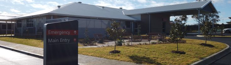 Main entrance to the Murrumburrah-Harden Health Service. A sign indicating the main and emergency entry points is shown to the left of the image. A curved bitumen driveway leads to the entry points. A part of a car park is shown on the left of the image. A concrete walkway runs along the topside of the car park. 